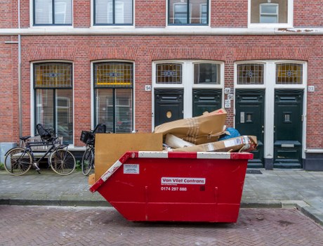 A large red skip bin positioned on a pavement in front of a red-brick residential building with three black front doors and tall windows. The bin contains various waste materials, including a large piece of cardboard and smaller items, with some debris visible spilling over the edges. To the left of the bin, two bicycles are parked and secured near the building's ground-floor windows, which have protective yellow bars. The scene is outdoors during daylight, with natural light illuminating the area, suggesting a typical urban setting suitable for rubbish removal services in Kings Langley or nearby areas. The image exemplifies waste collection in a residential environment, aligned with services offered by House Clearance Kings Langley.