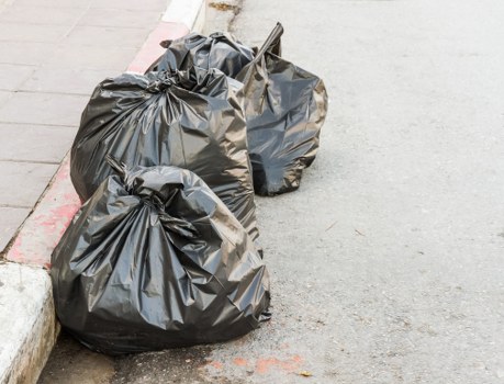 Two large black plastic rubbish bags filled with waste are positioned on a residential street pavement near the curb. The bags appear tightly packed and are placed close to the edge of the kerb, with one bag slightly leaning against the other. The surface underneath is concrete, showing minor cracks and some faint discoloration. To the left, a portion of a brick or stone curb edges the street, with a small strip of red-painted line marking the boundary between the pavement and the roadway, which appears to be made of asphalt. The environment suggests an outdoor setting typical of urban or suburban rubbish collection points, likely in a town such as Kings Langley. The scene is illuminated uniformly, indicating natural daylight, and the overall appearance points to a routine rubbish removal activity, possibly managed by a local waste management service such as House Clearance Kings Langley, operating within the postcode area inferred from the URL.