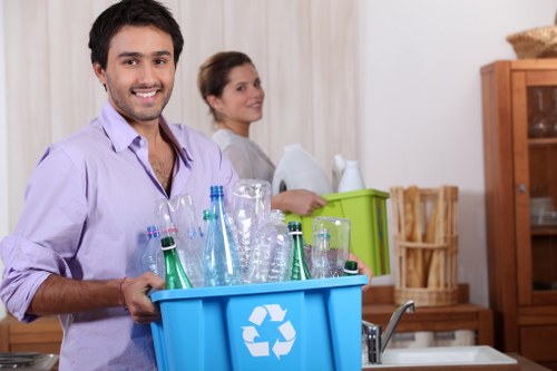 A smiling man with dark hair and a beard, dressed in a light purple shirt, holds a blue recycling bin filled with several glass bottles of varying shades of green and clear, and some transparent plastic containers, in a domestic kitchen setting. Behind him, a woman with brown hair tied back, wearing a light-colored top, stands near a green plastic box and a wooden kitchen cabinet, indicating an environment related to household waste sorting or rubbish collection. The background features wooden kitchen furniture, including a cupboard and a magazine rack, with neutral-toned curtains on the window, and the overall atmosphere is well-lit and tidy. The scene reflects typical rubbish removal or house clearance activities, with a focus on glass and plastic waste. House Clearance Kings Langley offers services related to rubbish collection and disposal, and the image subtly emphasizes environmentally responsible waste sorting at a residential property in the Kings Langley area, inferred from the context and setting.