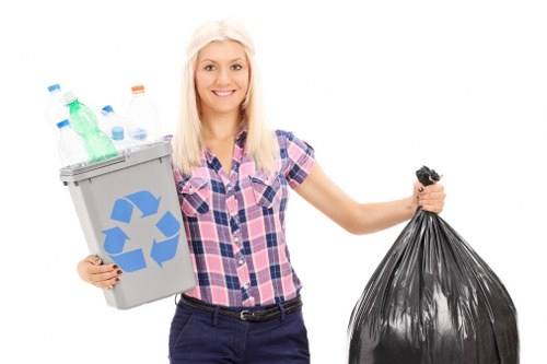 A young woman with long blonde hair, wearing a pink and blue checkered shirt and dark trousers, standing against a plain white background. She is smiling and holding a small grey recycling bin filled with various plastic bottles, including clear and green types, which are visibly crumpled or toppled over the edge of the bin. In her other hand, she carries a large black garbage bag made of plastic, appearing full and slightly wrinkled. The background is plain, emphasizing her expression and the waste collection items she is holding, illustrating a scene related to rubbish collection and responsible disposal. The overall image is clear, professional, and suitable for representing household rubbish removal services in the UK, especially around areas like Kings Langley.