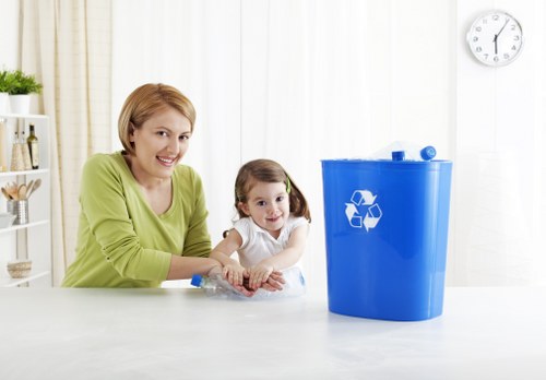 A woman with short, light brown hair and a young girl with dark blonde hair, both smiling, sit at a white table in a bright, modern room with light-colored curtains. The woman is wearing a light green long-sleeve top, and the girl is dressed in a white shirt. They are positioned next to a large, blue recycling bin with a white recycling symbol on the front, placed on the table. The room has a minimalistic decor, with a small shelf holding bottles and containers in the background, and a white wall with a round clock showing approximately 1:05. The scene appears to emphasize family involvement in environmentally conscious activities, relevant to household rubbish and waste management services, with a subtle connection to local services offered by House Clearance Kings Langley, near the postcode district of Kings Langley or surrounding areas in Hertfordshire.