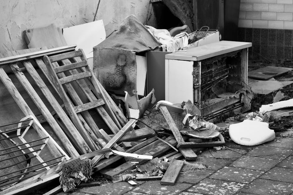 A collection of discarded household items and debris is piled on a paved outdoor surface, close to a brick wall and a wooden fence. The debris includes an overturned wooden garden trolley with slatted