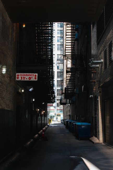 A narrow outdoor alleyway situated between a building with dark green horizontal metal siding on the left and a brick wall on the right, leading towards a background of leafless trees and a partly clo
