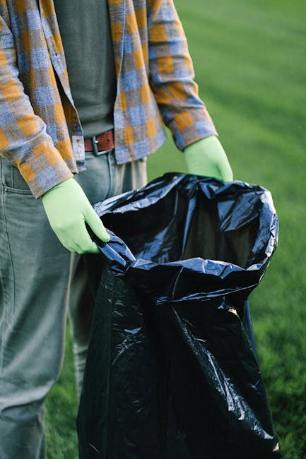 A person wearing a checkered yellow and gray shirt, light gray trousers, and bright green gloves is holding a large black rubbish bag open with both hands, preparing for waste collection. The bag is m