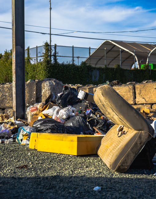 A person wearing a checkered yellow and gray shirt, light gray trousers, and bright green gloves is holding a large black rubbish bag open with both hands, preparing for waste collection. The bag is m