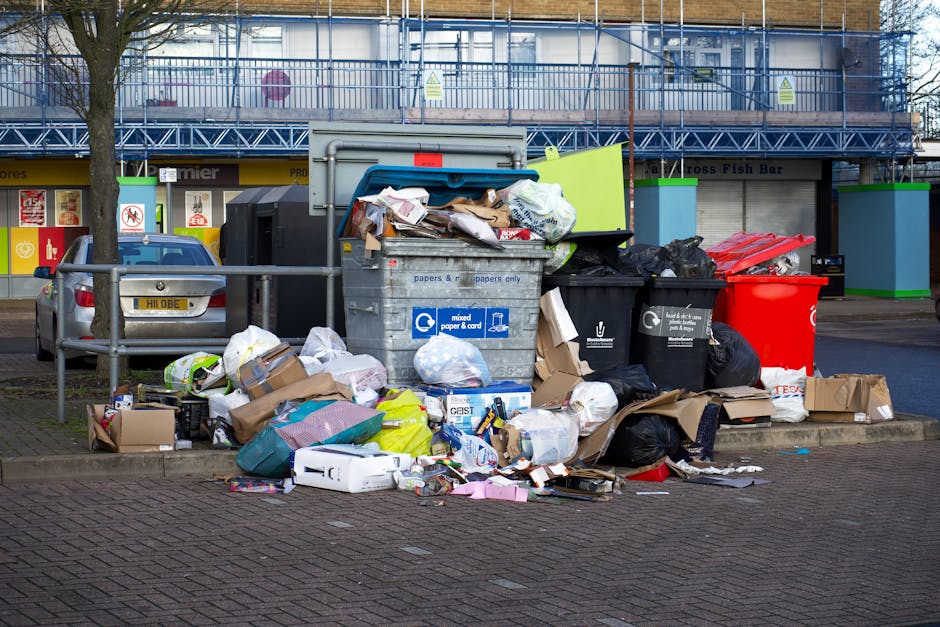 A person's hand is visible holding a green plastic rubbish bag by its twisted top, which is tied into a knot. The bag appears partially filled with waste, as indicated by its rounded shape and bulging
